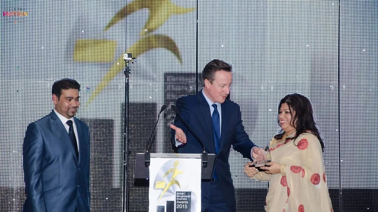A man in a suit standing at a podium handing an award to a woman in a sari, with another man in a suit standing to their left.